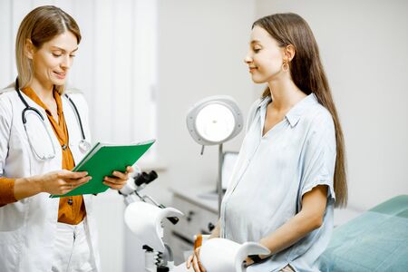 Young pregnant woman sitting on the gynecological chair during a medical consultation with gynecologist. Doctor writing medical history of a patientの写真素材