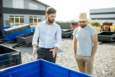 Agronomist with salesman choosing a new farm truck trailer, standing on the open ground of the...