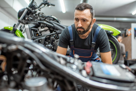 Portrait of a handsome bearded biker in working overalls sitting near the motorcycles during the repairment in the workshopの写真素材