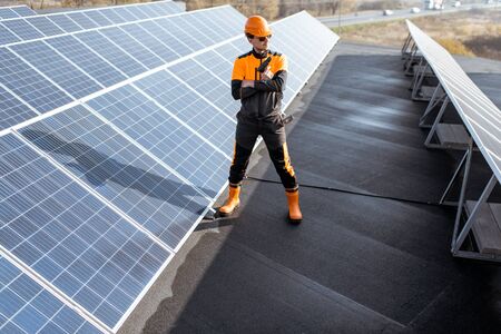 Portrait of a well-equipped worker in protective orange clothing examining solar panels on a photovoltaic rooftop plant. Concept of maintenance and installation of solar stationsの写真素材