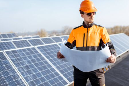 Portrait of an engineer in protective workwear with blueprints of solar station project on a photovoltaic rooftop plant. Concept of designing power plantsの写真素材