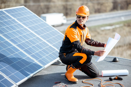 Electrician in protective workwear installing solar panels, sitting on a rooftop with blueprints and...