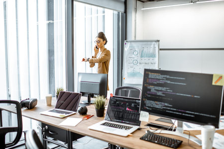 Young business woman talking on the phone while standing near the window in the modern office with...