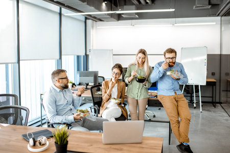 Group of diverse colleagues eating takeaway salad, sitting together and having fun during a...