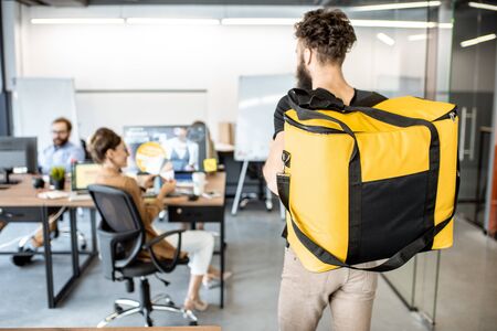 Courier delivering products for office workers, carrying yellow thermal bag, view from the back side. Concept of food delivery to employees in the officeの写真素材