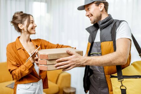 Handsome courier in uniform delivering pizza with thermal bag to a client home, happy woman receiving her orderの写真素材