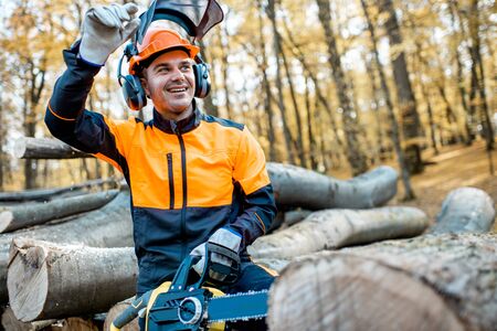 Portrait of a cheerful professional lumberjack in protective workwear standing with a chainsaw on a pile of logs in the forestの写真素材