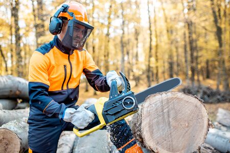 Professional lumberjack in protective workwear working with a chainsaw in the forest, sawing a thick wooden logの写真素材