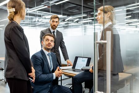 Group of strictly dressed business people having a discussion, sitting at the workplace with a computer in the modern office with glass partitionsの写真素材