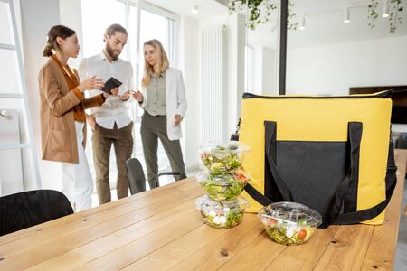 Office workers standing in the office with business lunches and delivery bag on the foreground. Concept of takeaway healthy food delivery to the workの写真素材