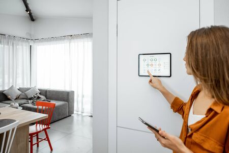 Young woman controlling home with a digital touch screen panel installed on the wall in the living room. Concept of a smart home and mobile application for managing smart devices at homeの写真素材