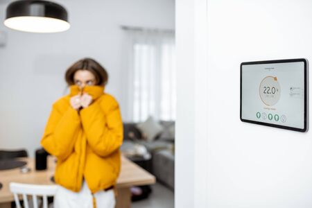 Digital panel for smart home managing with launched heating control application in the living room. Woman feeling cold on the backgroundの写真素材