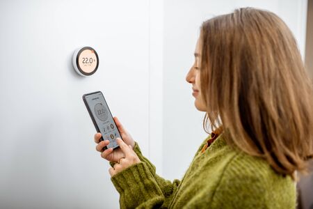 Woman dressed in green sweater regulating heating temperature with a modern wireless thermostat and smart phone at home. Synchronization of thermostat with mobile devices conceptの写真素材