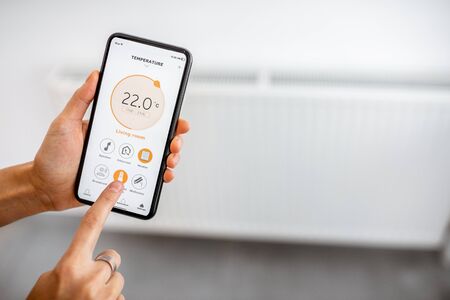 Controlling radiator heating temperature with a smart phone, close-up with radiator on the background. Concept of a smart home and mobile application for managing smart devices at homeの写真素材
