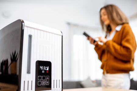 Close-up on a smart air humidifier with touch screen, woman controlling it with smart phone on the backgroundの写真素材