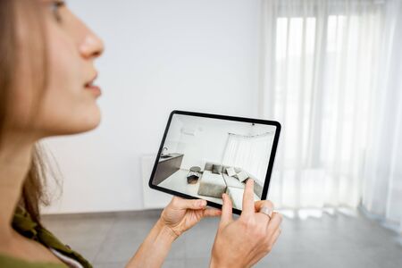 Young woman placing new furniture on a digital tablet into the empty interior, looking how it looks before buying. Concept of augmented reality in design and retail businessの写真素材