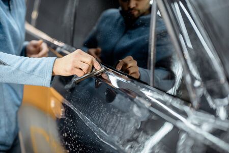 Worker sticking anti-gravel film on a car body with scrapper at the detailing vehicle workshop, close-up. Concept of car body protection with special filmsの写真素材