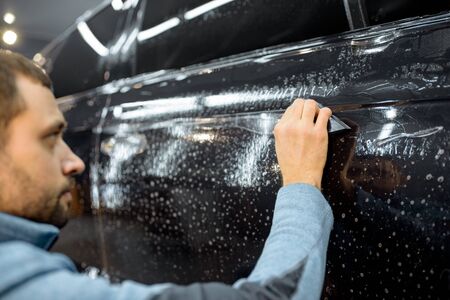 Car service worker sticking anti-gravel film on a car body with scrapper at the detailing vehicle workshop. Concept of car body protection with special filmsの写真素材