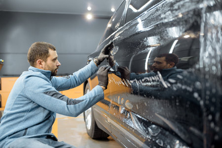 Car service worker sticking anti-gravel film on a car body with scrapper at the detailing vehicle workshop. Concept of car body protection with special filmsの写真素材