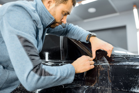 Car service worker sticking anti-gravel film on a car body with scrapper at the detailing vehicle workshop. Concept of car body protection with special filmsの写真素材