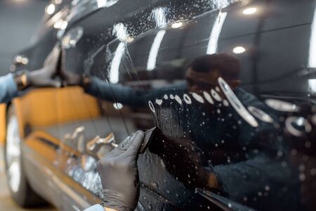 Worker sticking anti-gravel film on a car body with scrapper at the detailing vehicle workshop, close-up. Concept of car body protection with special filmsの写真素材