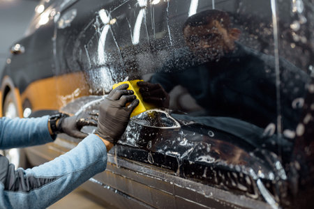 Worker sticking anti-gravel film on a car body with scrapper at the detailing vehicle workshop,...