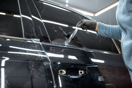 Worker trimming with removing remains of a protective film, sticking it on a car body at the vehicle service, close-up. Concept of car body protection with special filmsの写真素材
