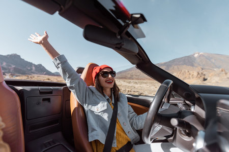 Happy woman in red hat driving convertible car while traveling on the desert road. Carefree...