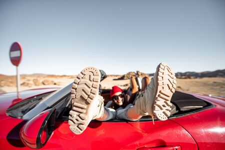 Woman traveling on converrtible sports car, pulling legs out the window. Side view on the red car with trekking shoesの写真素材