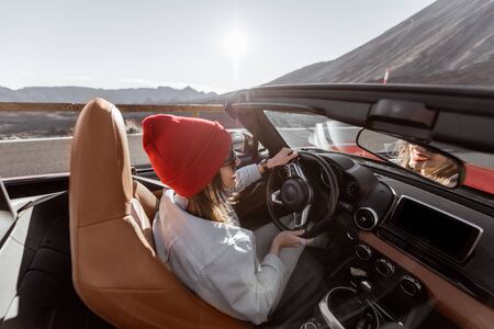 Happy woman in red hat driving convertible car while traveling on the desert road. Carefree lifestyle and travel conceptの写真素材