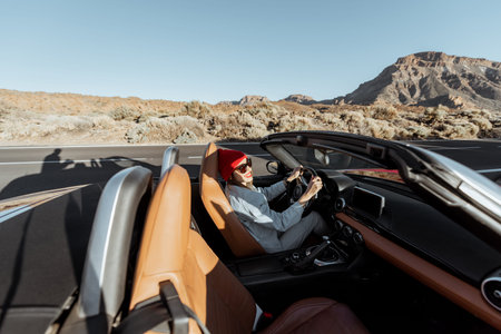 Happy woman in red hat driving convertible car while traveling on the desert road. Wide view from...
