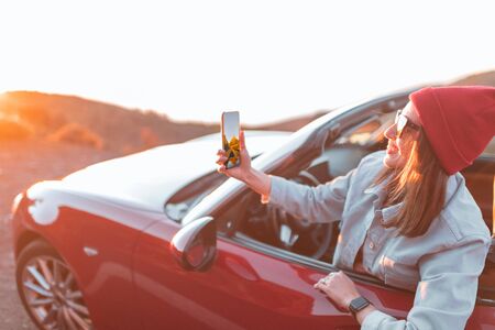Young woman enjoying beautiful sunset view, leaning out of the car window with mobile phone. Carefree lifestyle and travel conceptの写真素材