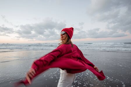 Lifestyle portrait of a carefree woman dressed in red shirt and hat walking on the beach at dusk. Wellness, happiness and life enjoyment conceptの写真素材
