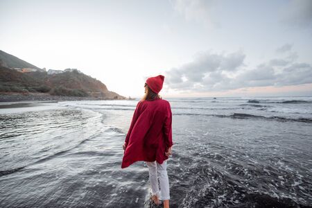 Lifestyle portrait of a carefree woman dressed in red shirt and hat walking on the beach at dusk. Wellness, happiness and life enjoyment conceptの写真素材
