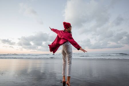 Lifestyle portrait of a carefree woman dressed in red shirt and hat walking on the beach at dusk. Wellness, happiness and life enjoyment conceptの写真素材