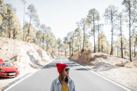 Lifestyle portrait of a carefree woman standing on the middle of the mountain road, with car on the backgroundの写真素材