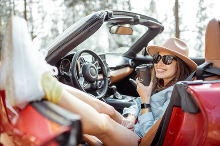 Young stylish woman enjoying traveling by convertible car, sitting freely with cofffee drink on the...