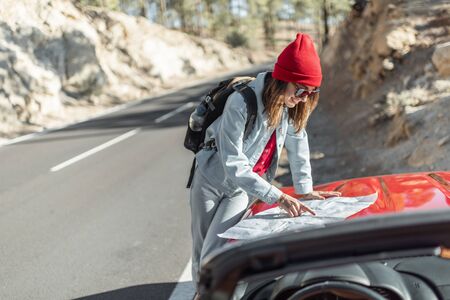 Young woman traveling by car in the mountains, having a break, standing with map on the roadsideの写真素材