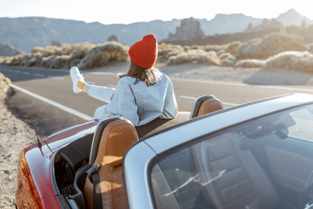 Woman enjoying beautiful rocky landscapes, sitting on the car hood on the roadside during a sunset....