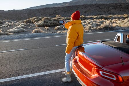 Woman enjoying beautiful rocky landscapes, photographing on phone near a car on the desert roadside during a sunset. Traveling on the volcanic valley on Tenerife islandの写真素材