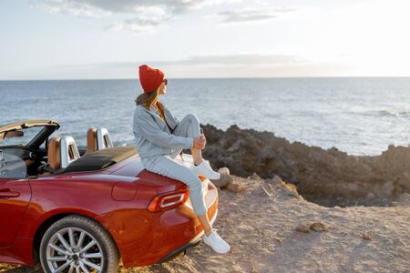 Woman enjoying beautiful view on the ocean, sitting on the convertible car on the rocky coast during a sunset. Carefree lifestyle and car travel conceptの写真素材