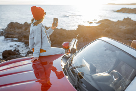 Young stylish woman enjoying beautiful landscapes, sitting with a coffee drink while travel on the rocky coastline during a sunset. Coffee enjoyment and car travel conceptの写真素材