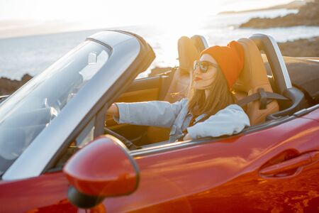 Lifestyle portrait of happy woman in red hat driving convertible car on the rocky ocean coast during a sunset. Carefree lifestyle and car travel conceptの写真素材
