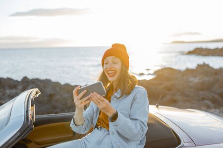 Young stylish woman photographing or vlogging on phone while traveling by car on the rocky coast near the ocean. Lifestyle travel and social influencing conceptの写真素材