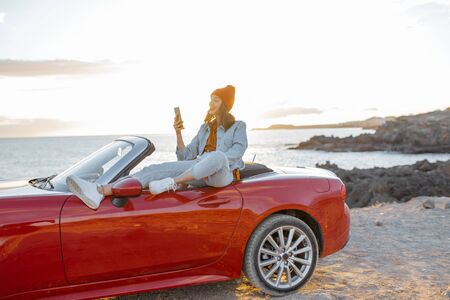 Young stylish woman photographing or vlogging on phone while traveling by car on the rocky coast near the ocean. Lifestyle travel and social influencing conceptの写真素材