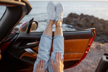 Woman traveling on car near the ocean, pulling legs out of the car, close-up on legs in white shoes with ocean landscape on the background. Carefree travel and nature enjoyment conceptの写真素材