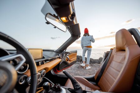 Woman enjoying beautiful view on the ocean, standing on the rocky coast during a sunset, view from the vehicle interior. Nature enjoyment and car travel conceptの写真素材