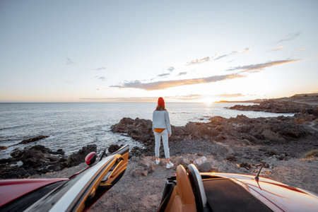 Woman enjoying beautiful view on the ocean, standing on the rocky coast during a sunset, view from the vehicle side. Nature enjoyment and car travel conceptの写真素材