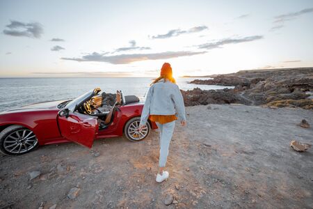 Pretty woman in red hat walking on the rocky coast straight to the ocean, traveling by a cabriolet during a sunsetの写真素材
