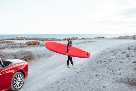Portrait of a young woman surfer in swimsuit walking with surfboard on the dirt road near the ocean. Active lifestyle and surfing conceptの写真素材